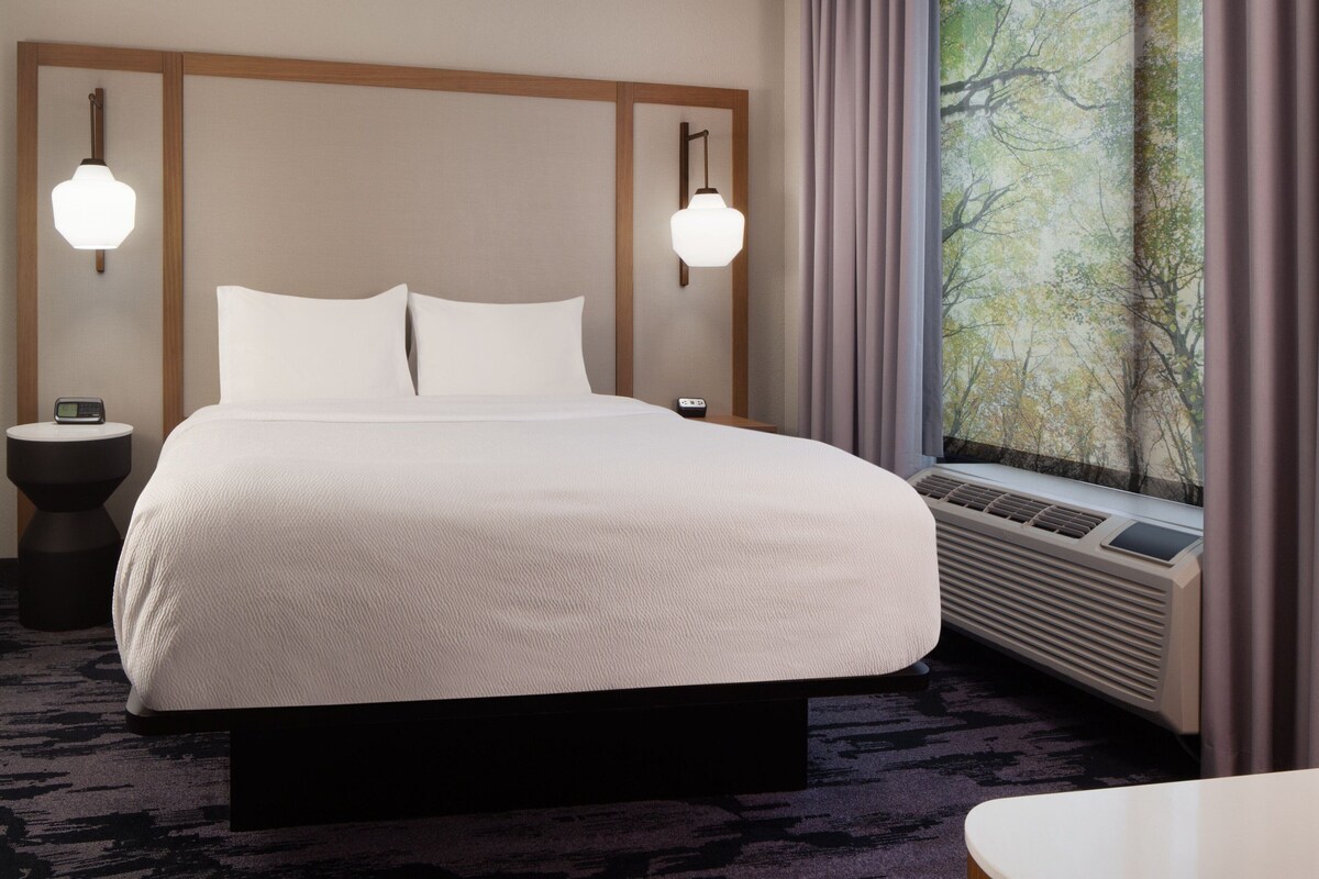 Habitación de hotel con una cama grande, dos mesitas de noche, un radiador montado en la pared y un ventanal con cortinas.