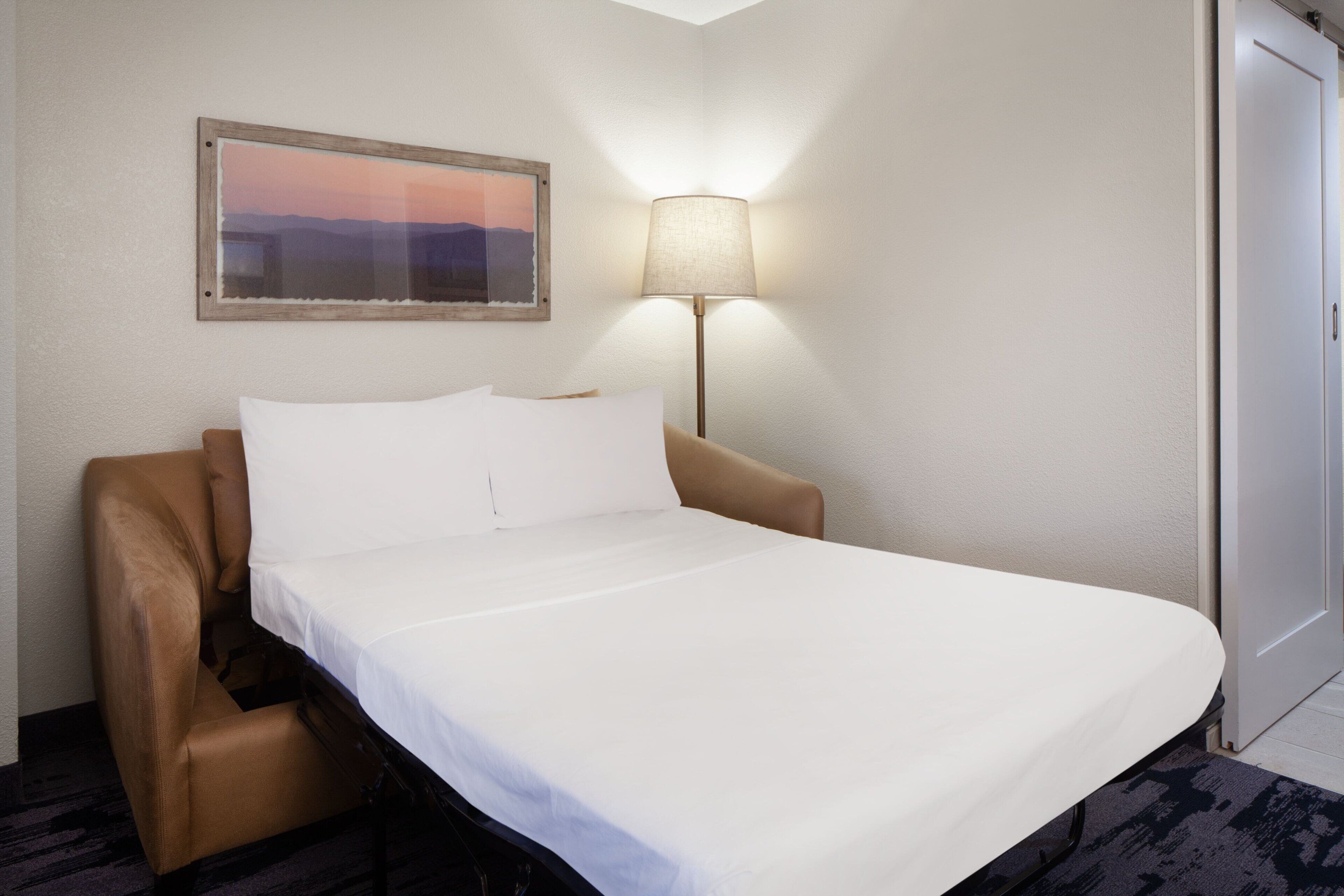 A hotel room with a bed, a chair, a lamp, a door, and a framed picture on the wall.