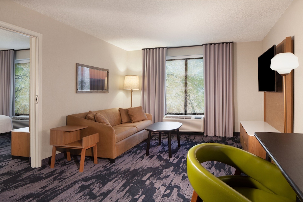 A hotel room with a sofa, a small table, a green chair, a lamp, a TV, and a window with curtains.