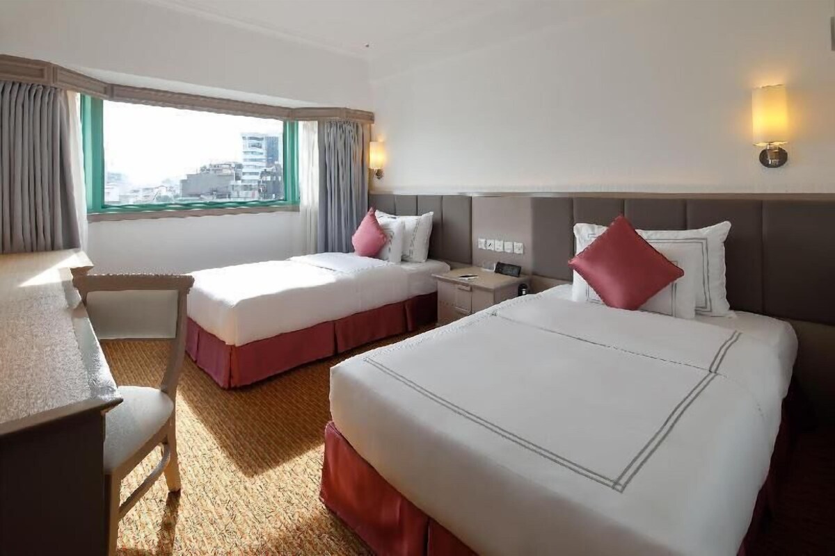 Une chambre d’hôtel avec deux lits, un bureau, une chaise, une fenêtre avec des rideaux et une table de chevet avec une lampe.