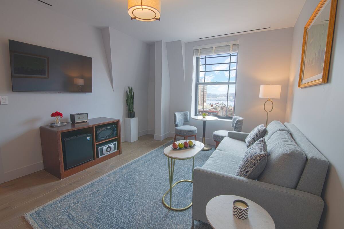 Una sala de estar moderna con un sofá gris, una mesita redonda, un televisor montado en la pared y una ventana con vistas a la ciudad.