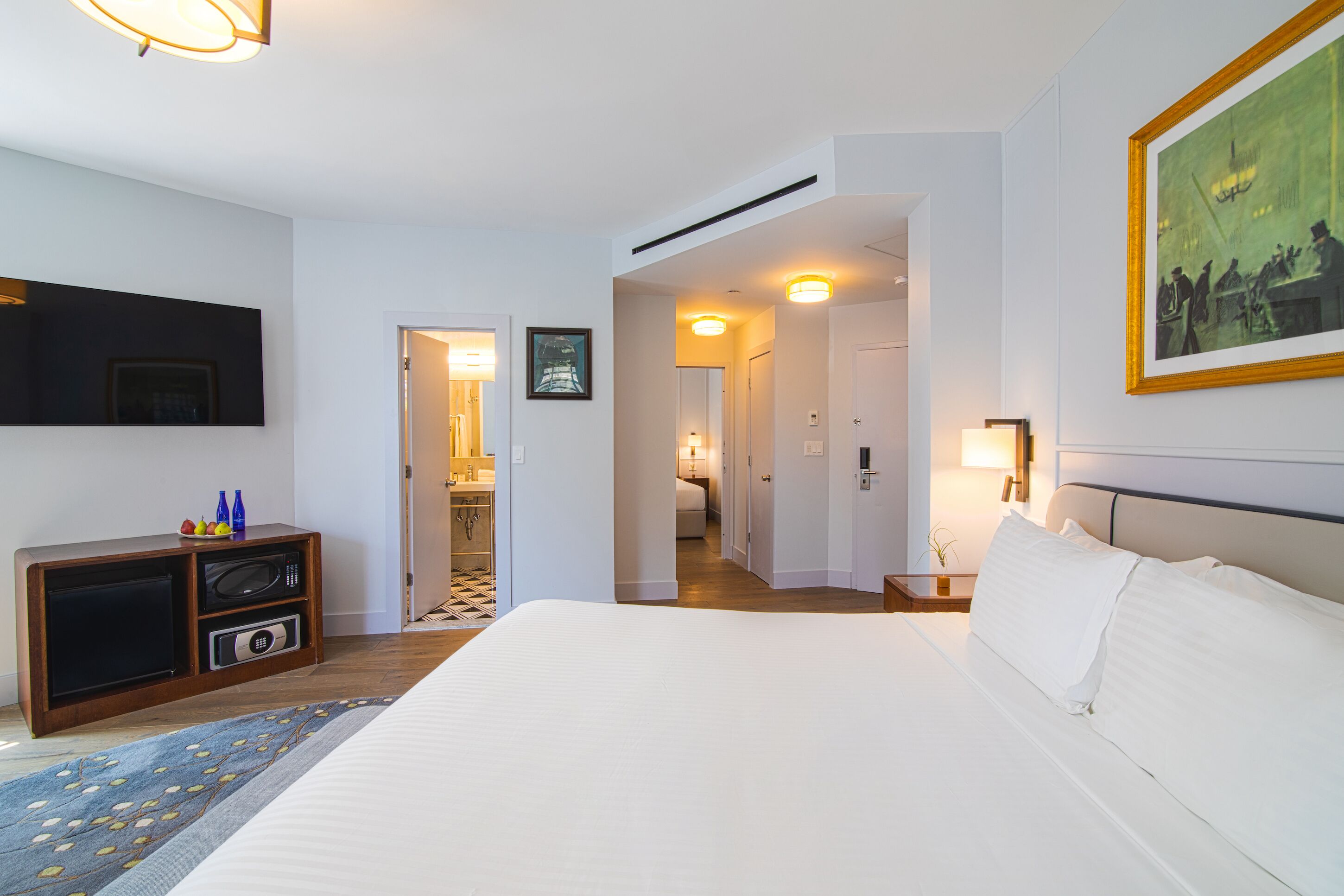 A hotel room with a large bed, a flat-screen TV, a microwave, and a small cabinet. There is a door leading to a bathroom and a hallway.