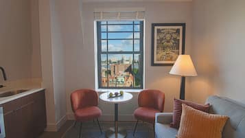 Une pièce compacte comprenant un coin cuisine, une table à manger avec deux chaises, un canapé avec des coussins, une fenêtre donnant sur la ville et une lampe.