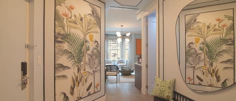 Un pasillo con ilustraciones botánicas enmarcadas en las paredes, un banco con cojín estampado, y una vista hacia una sala de estar con sofá y mesa de centro.