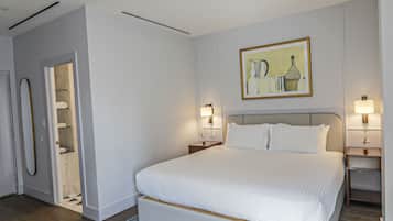 Una habitación de hotel moderna con una cama grande, mesitas de noche con lámparas, un cuadro en la pared y un baño visible a través de una puerta abierta.