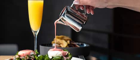 Una mano vertiendo una salsa rosa sobre un plato con verduras y un huevo escalfado, con un vaso de jugo de naranja al fondo.