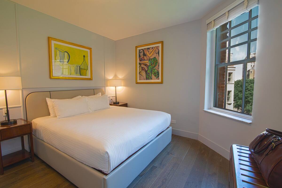 Una cama bien tendida en una habitación con dos mesitas de noche, cada una con una lámpara. Hay dos obras de arte enmarcadas en las paredes y una ventana con vistas a edificios y árboles.