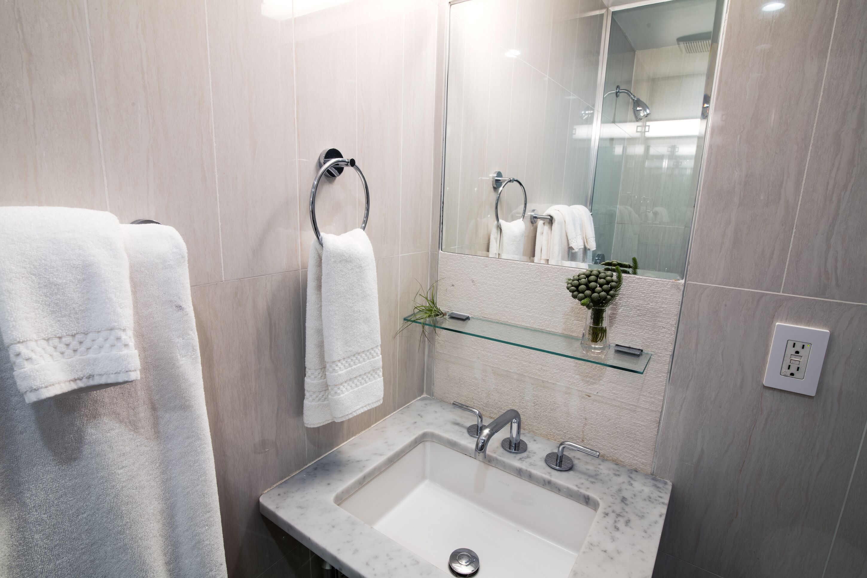 A modern bathroom with a marble sink, two towel rings, a glass shelf with a vase, and a large mirror.