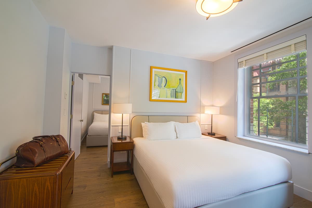 Habitación de hotel con una cama grande, lámparas de noche, un aparador de madera, un cuadro en la pared y una ventana con vistas a la vegetación.