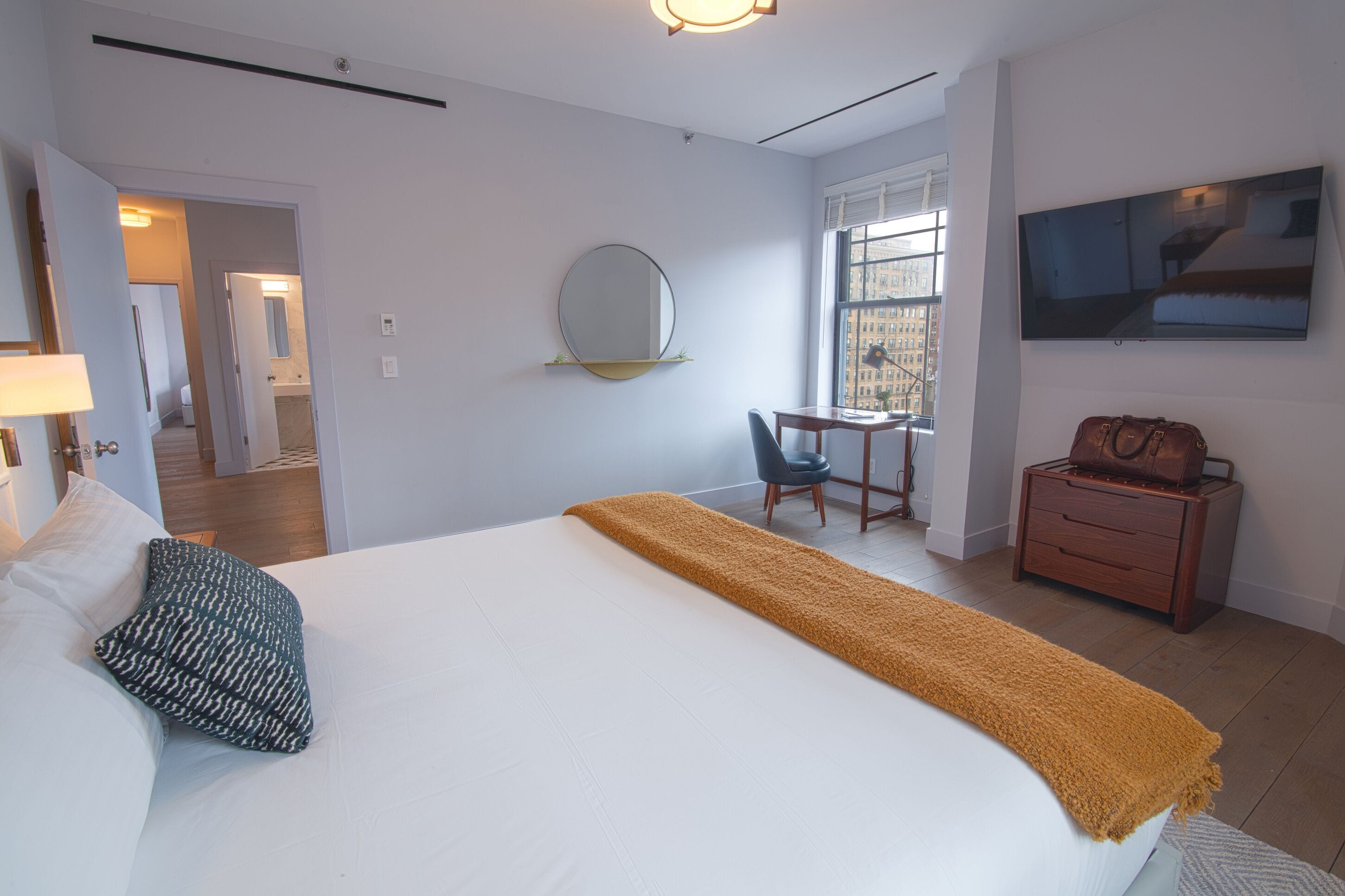 A hotel room with a bed, a desk, a chair, a mirror, a TV, and a suitcase.