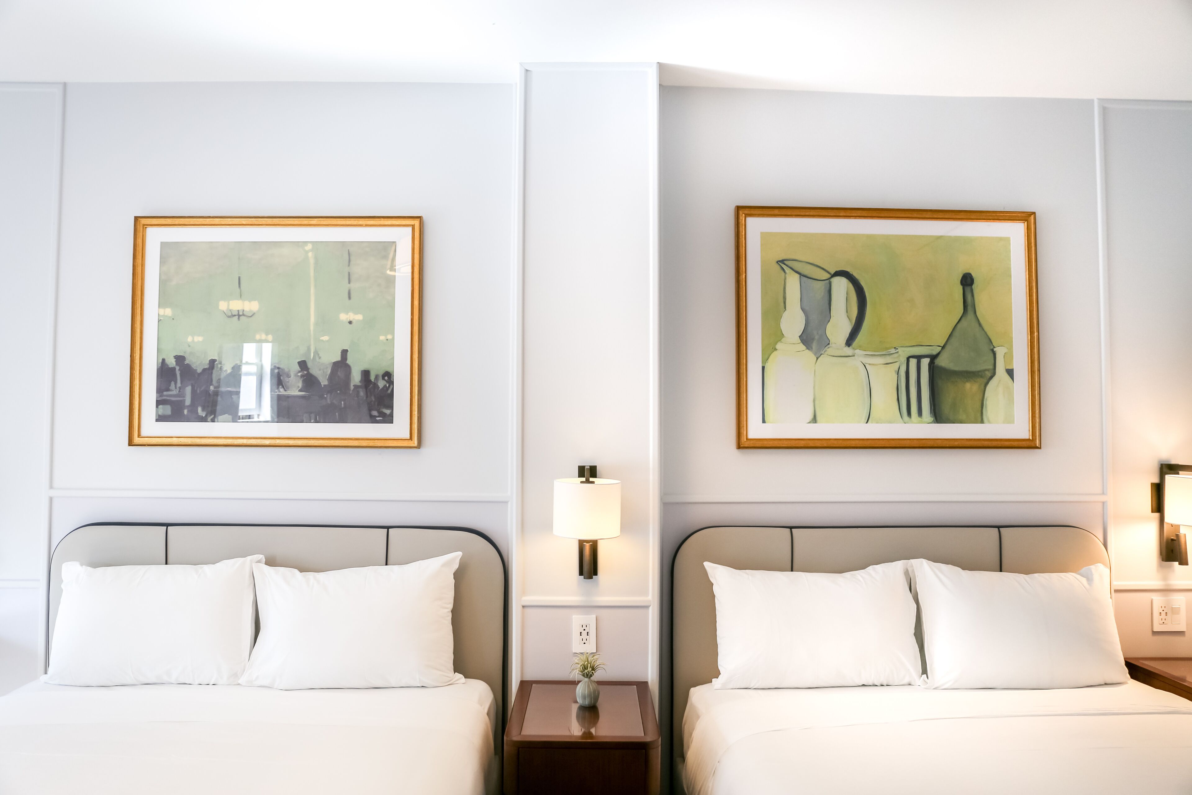Ein Hotelzimmer mit zwei Betten, einem Nachttisch mit Lampe und zwei gerahmten Kunstwerken an der Wand.