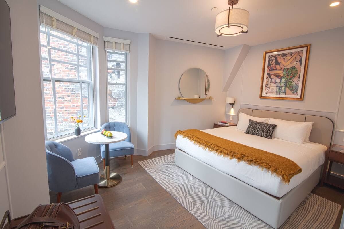 Una habitación de hotel moderna con una cama grande, un espejo redondo, una mesita pequeña y una silla azul.
