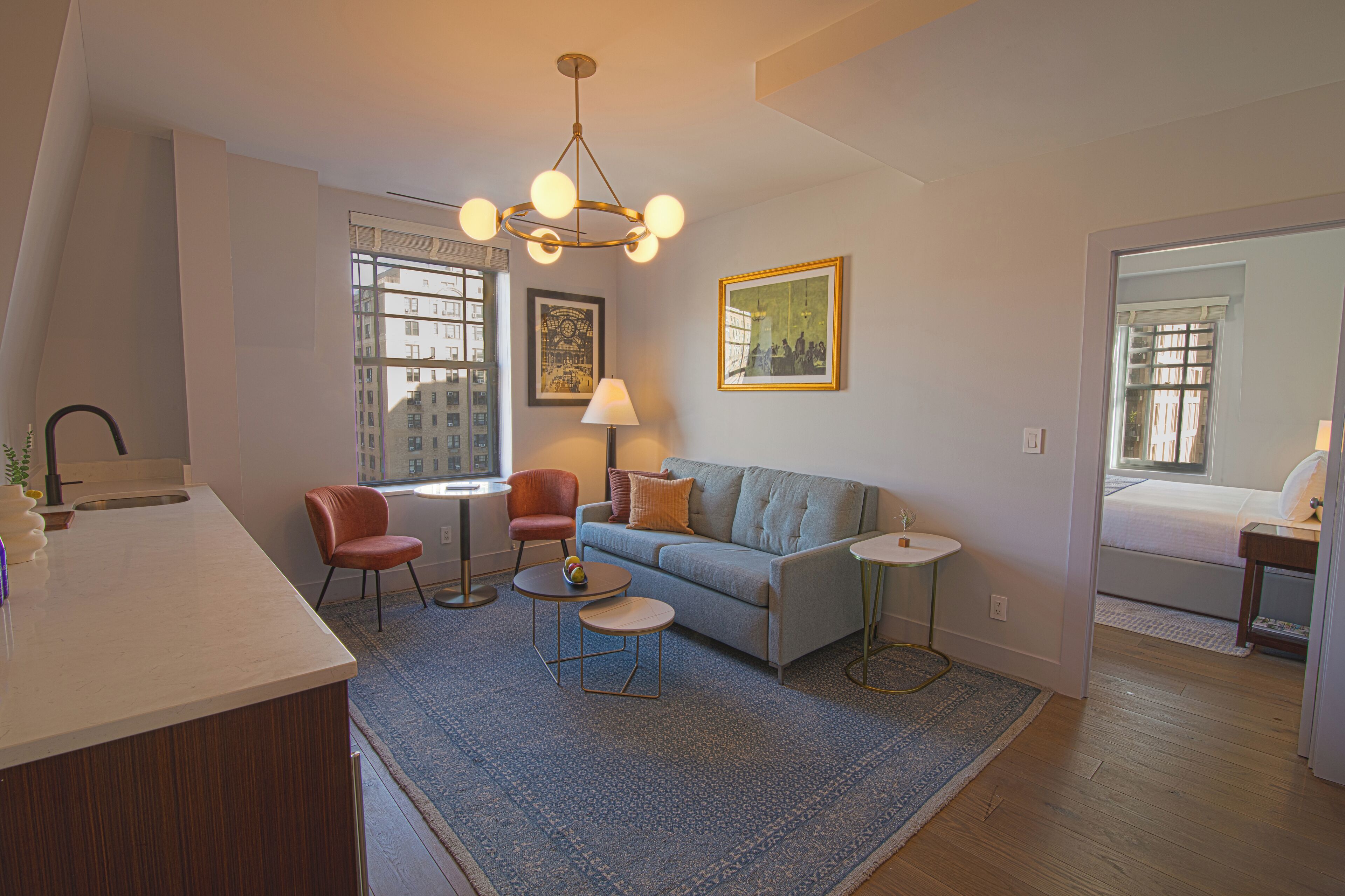 A modern living room with a sofa, two chairs, a small table, and a lamp. There is a window with blinds, a painting on the wall, and a door leading to another room.