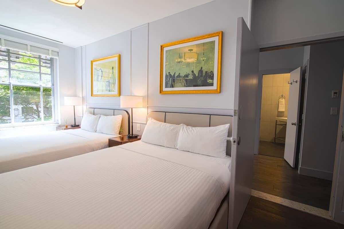 Habitación de hotel con dos camas, un ventanal grande y una puerta que da a un baño.
