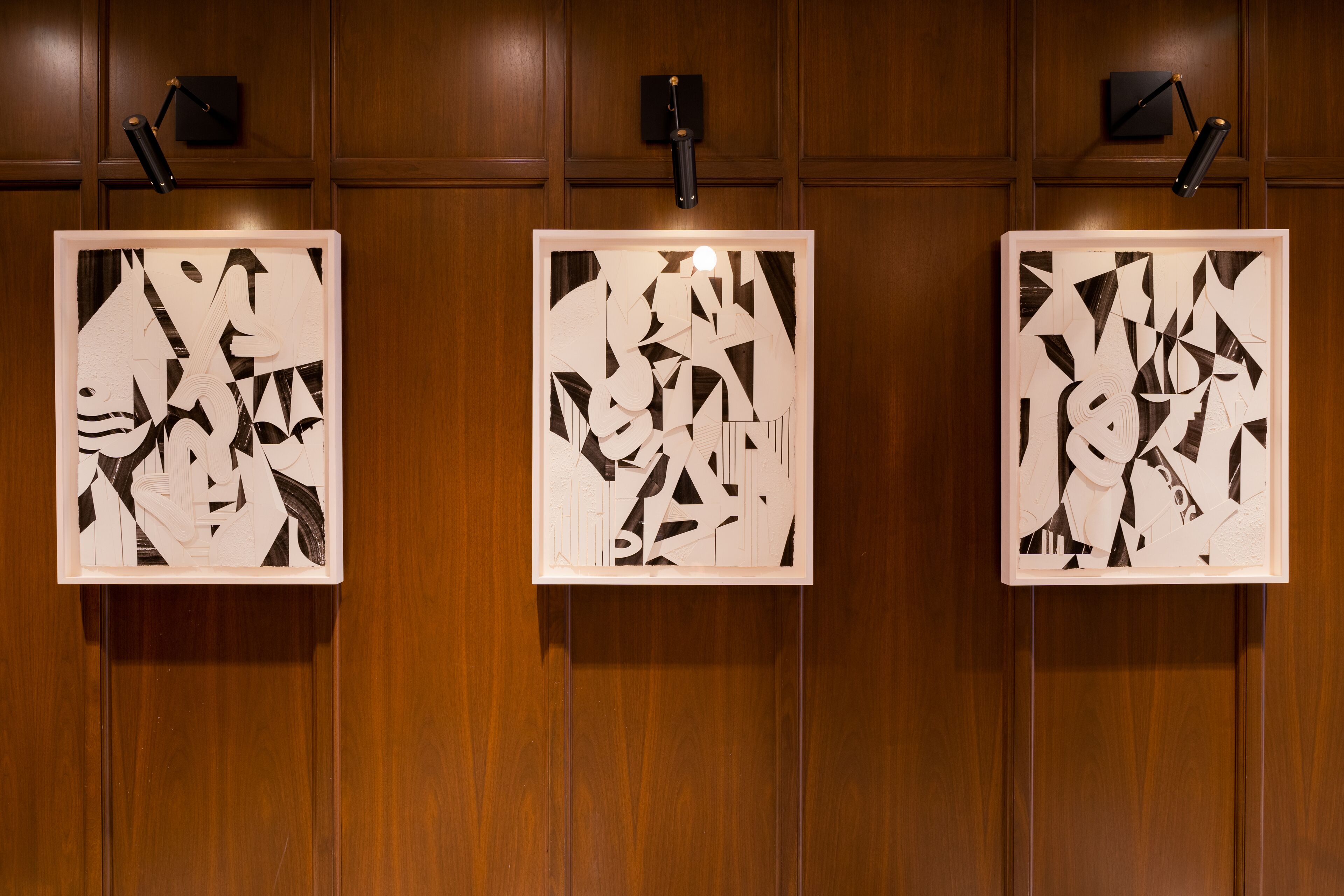 Tres obras de arte abstracto enmarcadas sobre una pared de madera con iluminación empotrada.