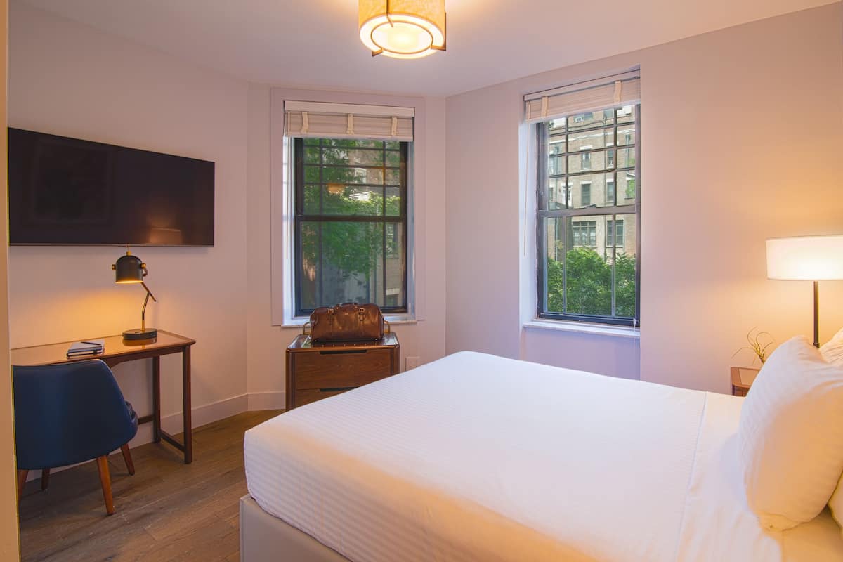 Un dormitorio con cama, escritorio, silla, lámpara y dos ventanas.
