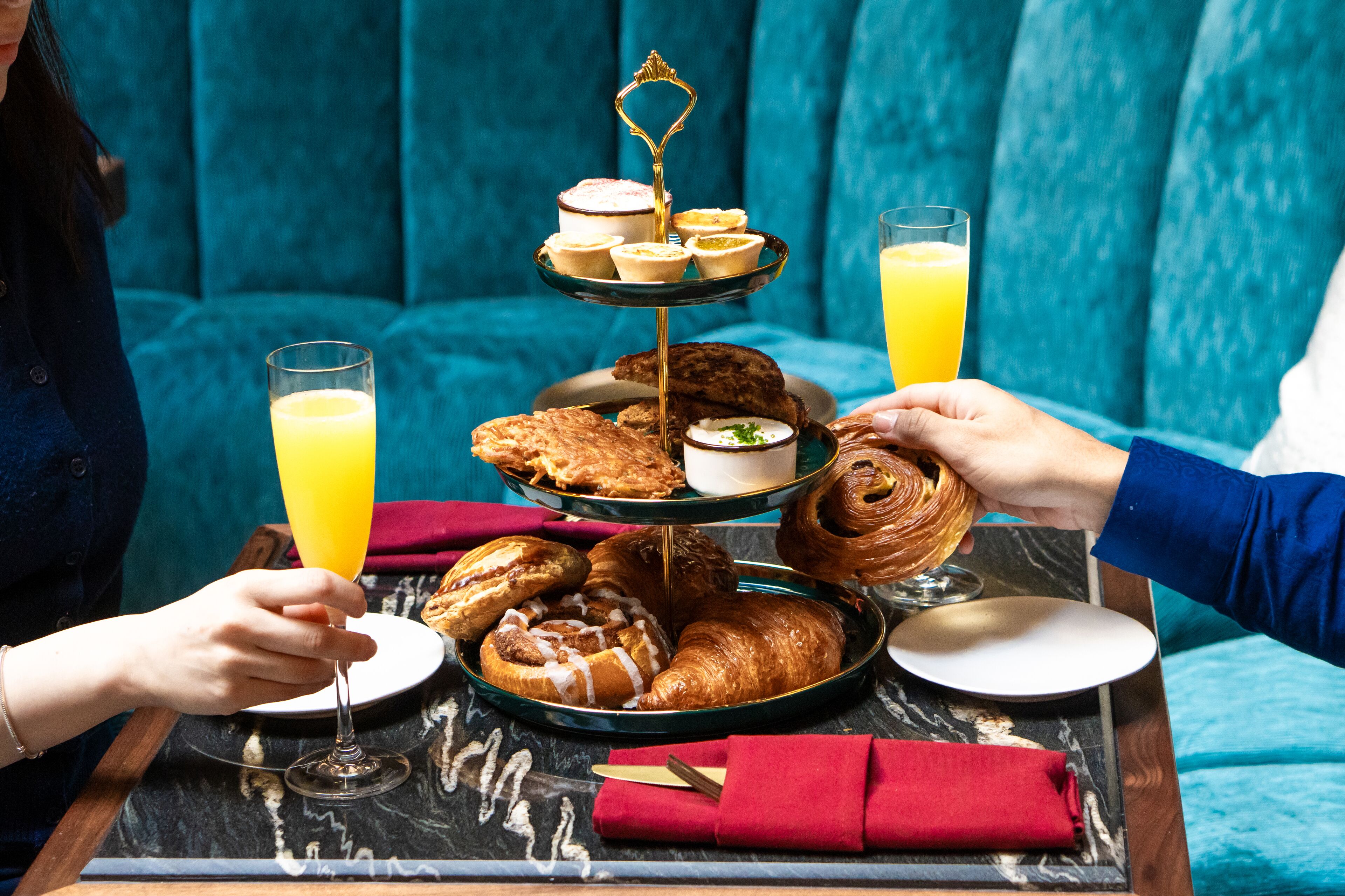 Una bandeja de desayuno con pasteles, un pastelero escalonado y vasos de zumo de naranja sobre una mesa de mármol.