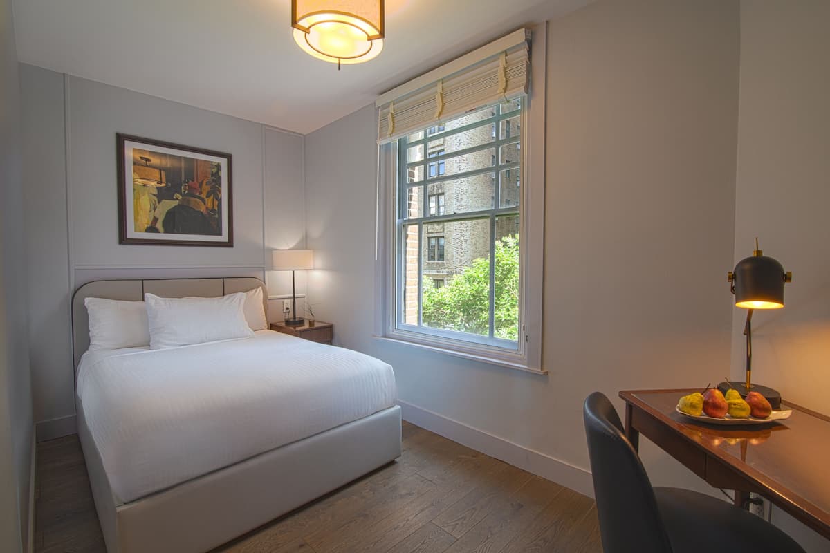 Habitación de hotel con cama, escritorio con lámpara, un plato de fruta, un cuadro en la pared y una ventana con vista a follaje.