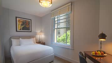 A hotel room with a bed, a desk with a lamp, a plate of fruit, a painting on the wall, and a window with a view of greenery.