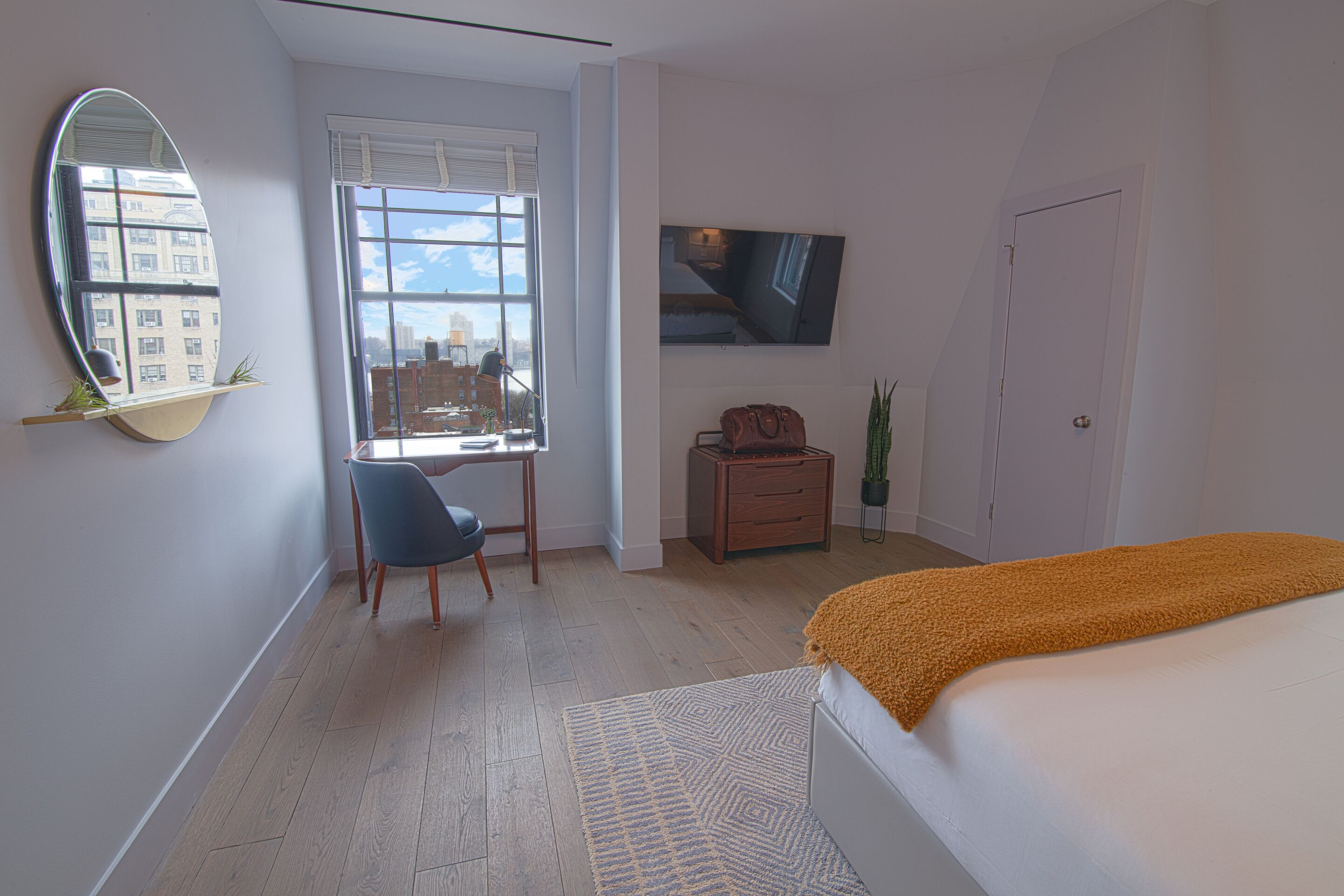 A bedroom with a bed, a desk, a chair, a TV, and a window with a city view.