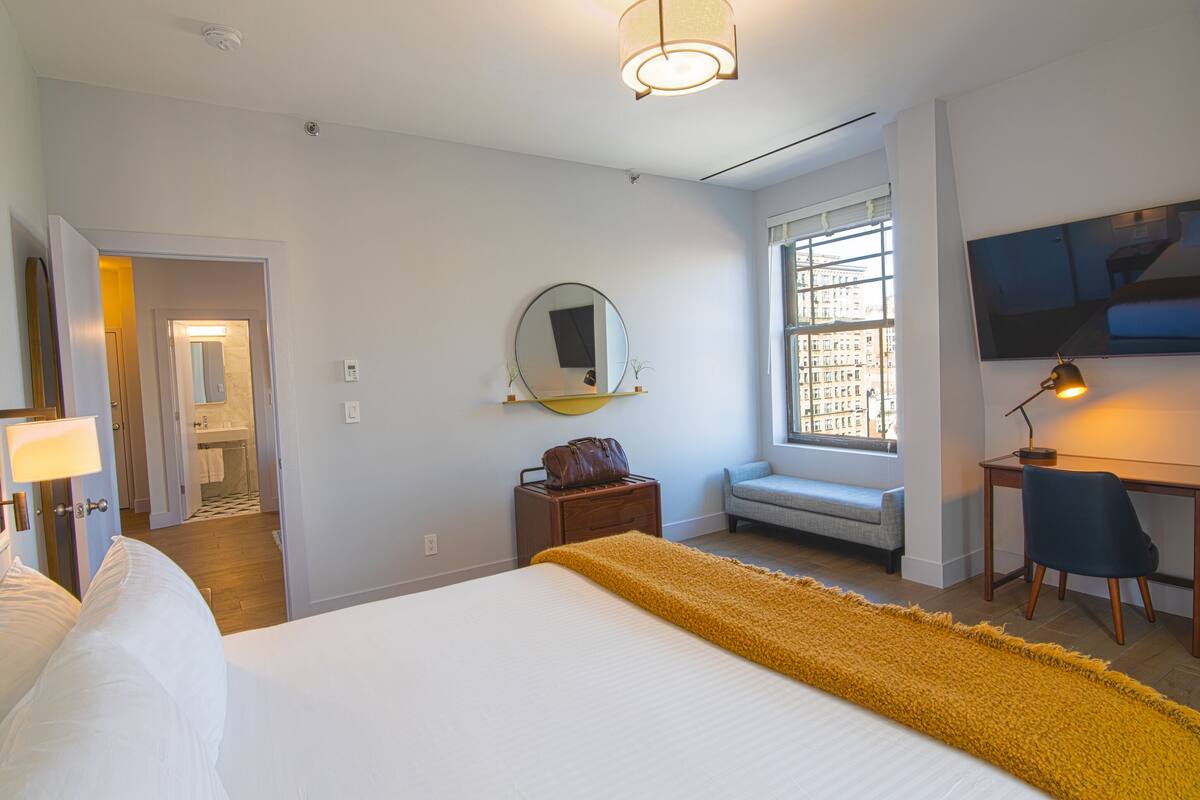 Una habitación de hotel con cama, escritorio, silla, ventana, espejo y baño a la vista a través de una puerta abierta.