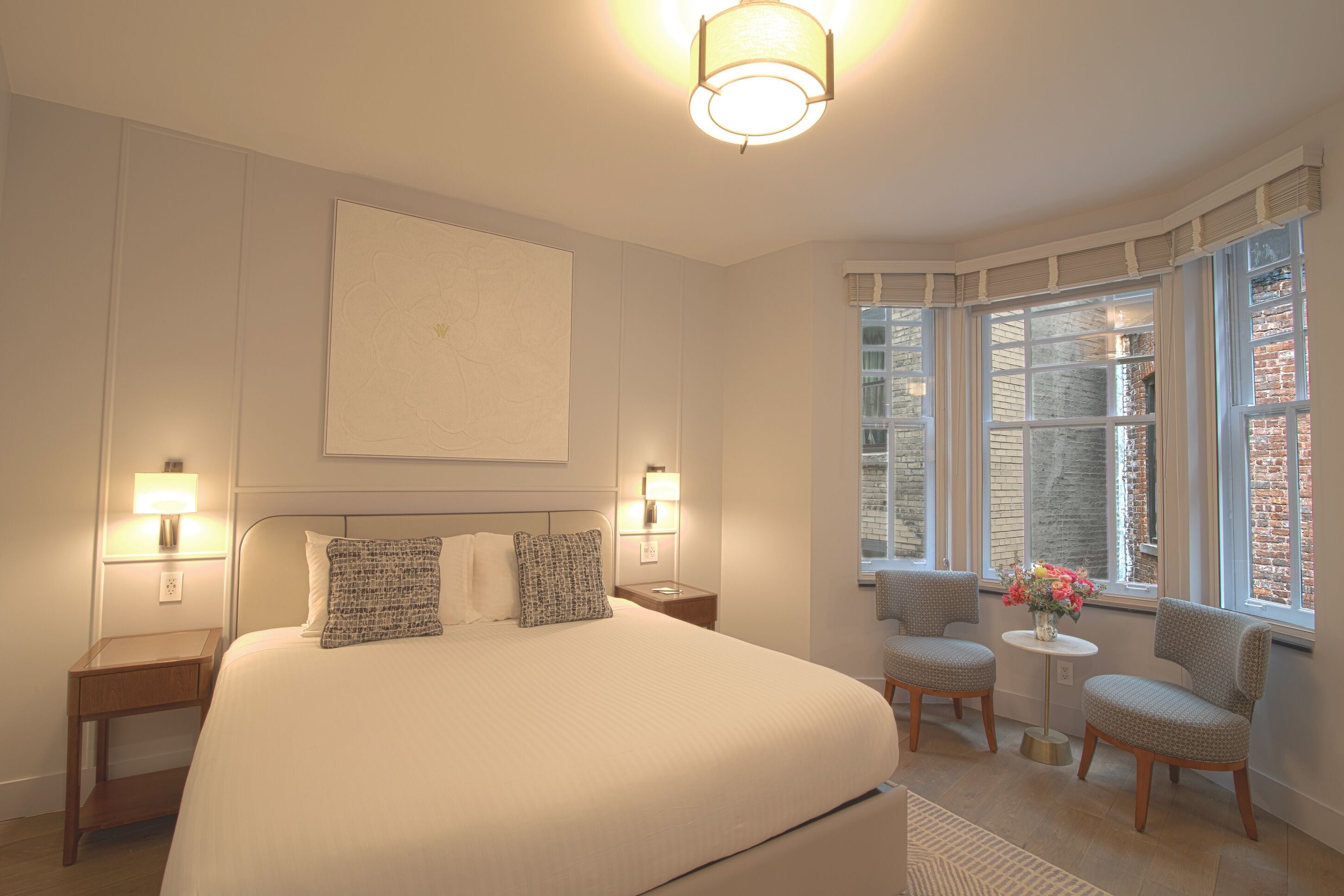 A hotel room with a large bed, two bedside lamps, a small table with chairs, and a view of brick buildings through large windows.