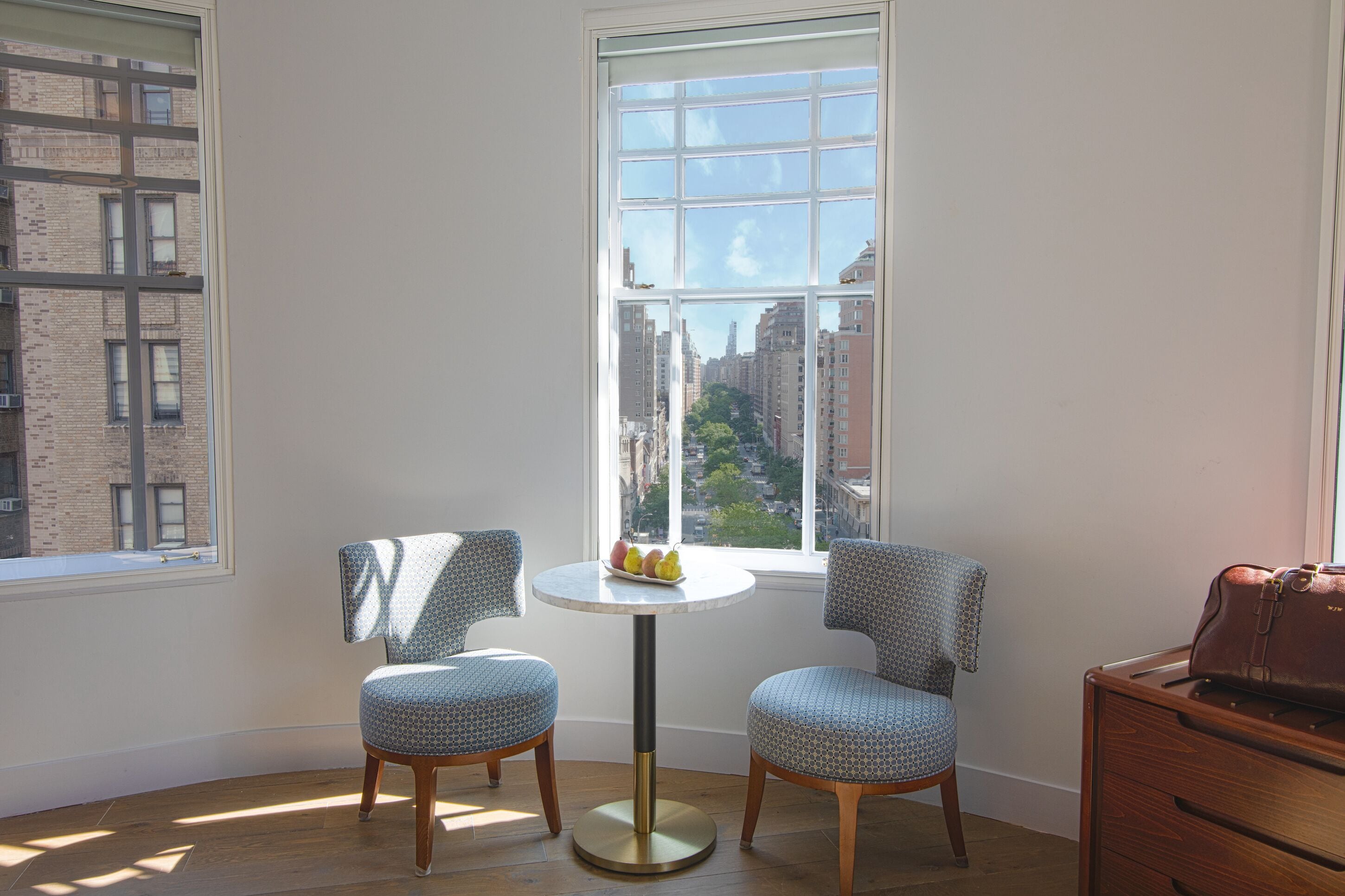 Habitación de esquina con dos sillones estampados, una mesita con fruta y vistas a un paisaje urbano a través de un ventanal grande.