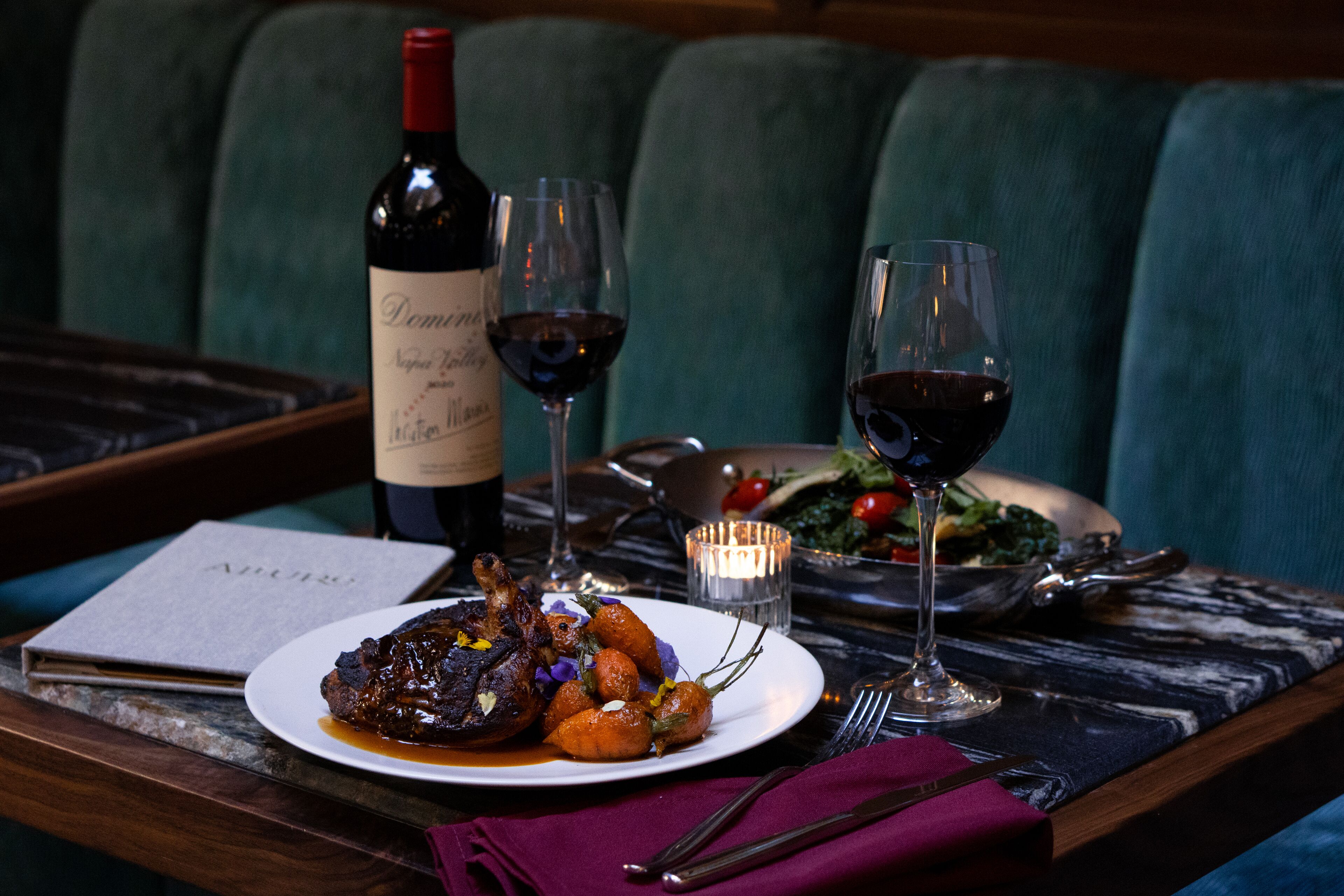 Una mesa de comedor con un plato de comida, dos copas de vino, una botella de vino y un menú.