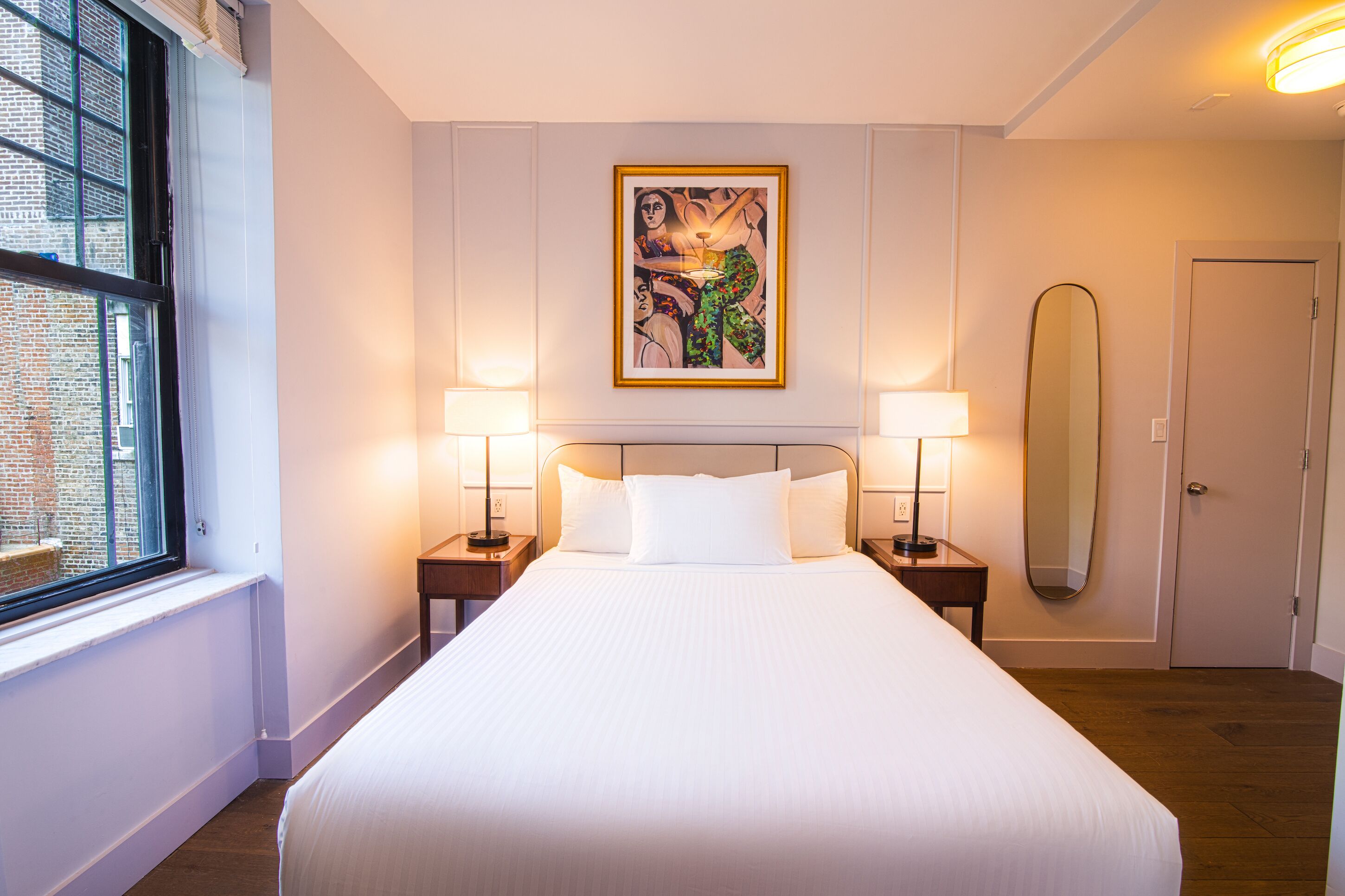 Habitación de hotel con una cama grande, dos lámparas de noche, un cuadro en la pared, un espejo y una ventana con vista a un edificio de ladrillo.