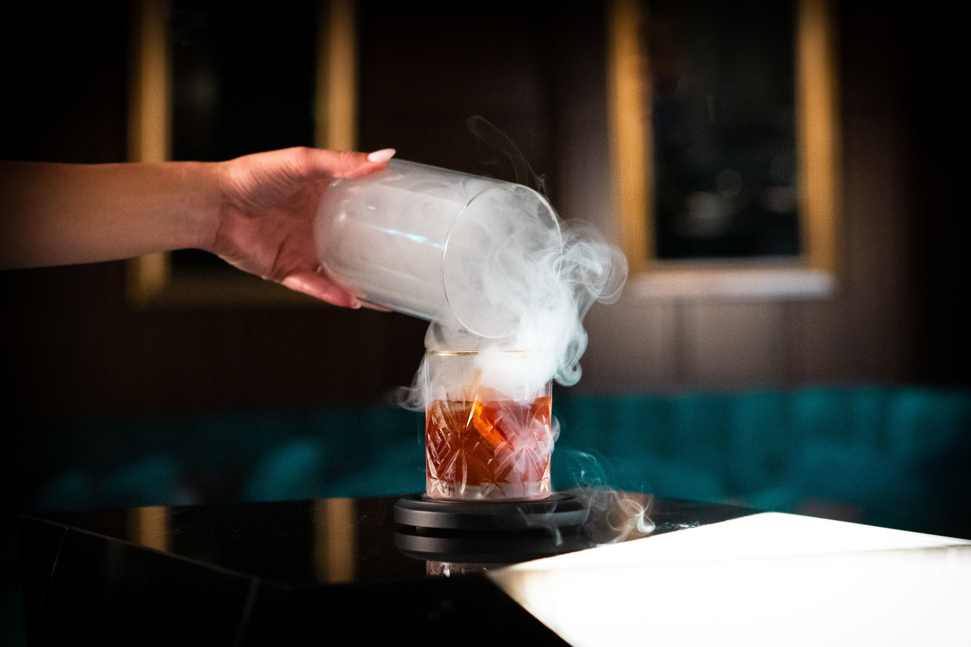 A hand pouring a clear liquid into a glass, creating a smoky effect.
