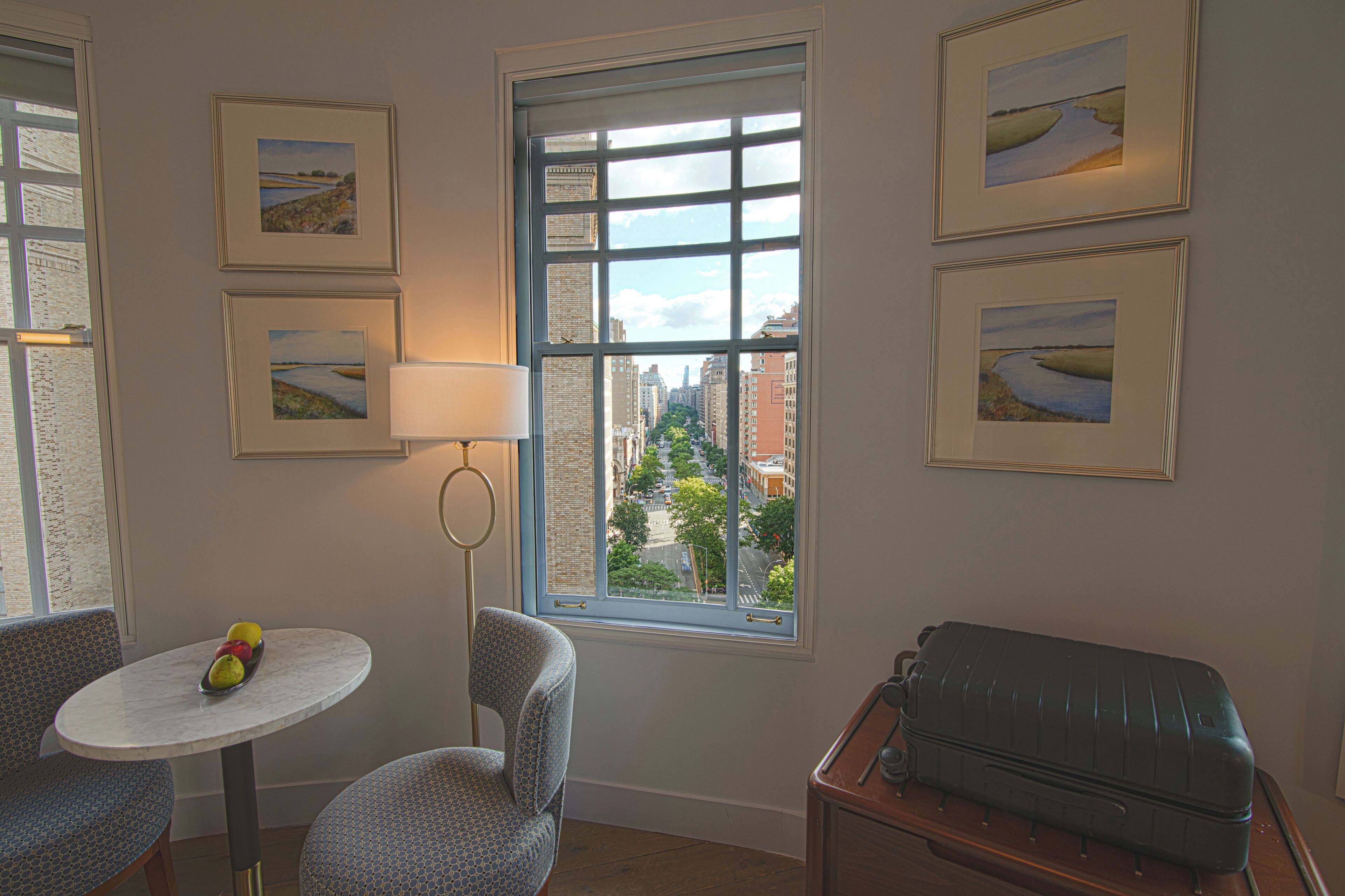 A corner room with a window, a table with fruit, a chair, and a suitcase.