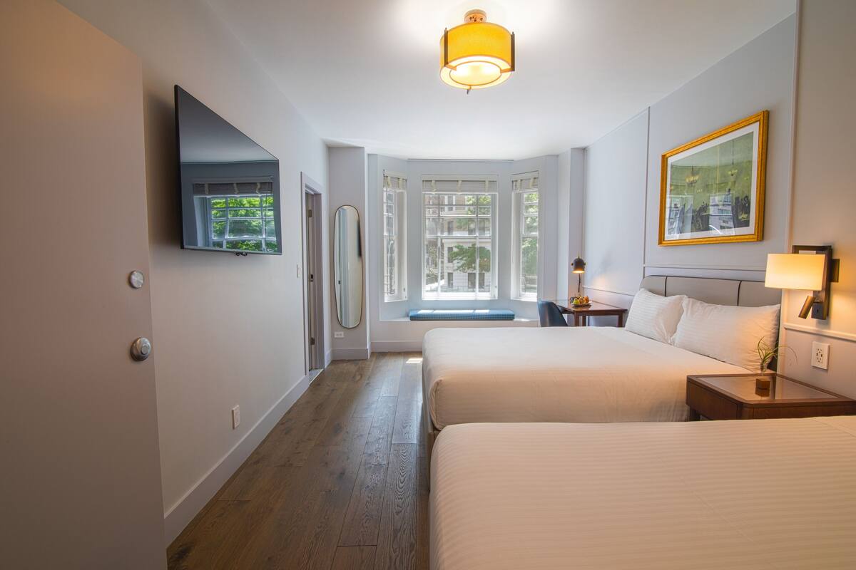 Habitación de hotel con dos camas, un televisor de pantalla plana montado en la pared, una ventana con vistas a zonas verdes y una mesita de noche con lámpara.