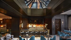 A modern bar area with a high ceiling and skylight, featuring a bar counter with numerous bottles, seating with blue upholstered chairs, and a dark wood finish.