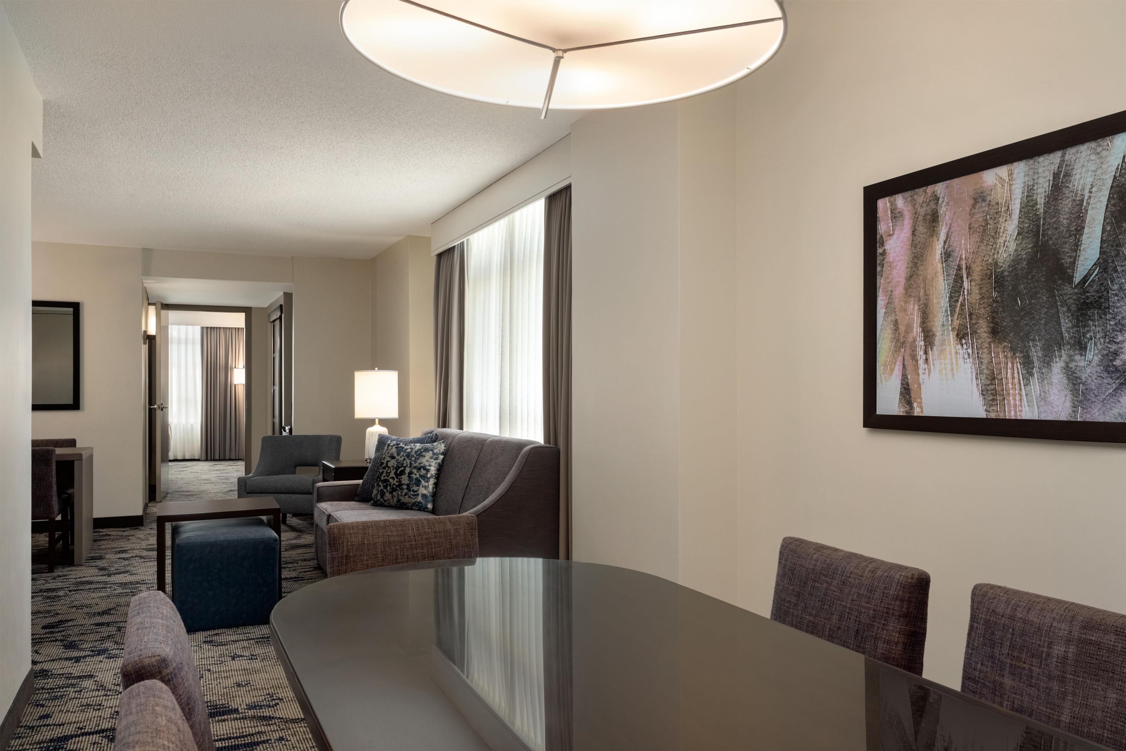 A hotel room with a large dining table, a sofa, a chair, a lamp, and a painting on the wall.