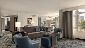 Habitación de hotel con sofá, sillones, una mesa de centro, televisor, lámpara, un cuadro y vistas a la ciudad.