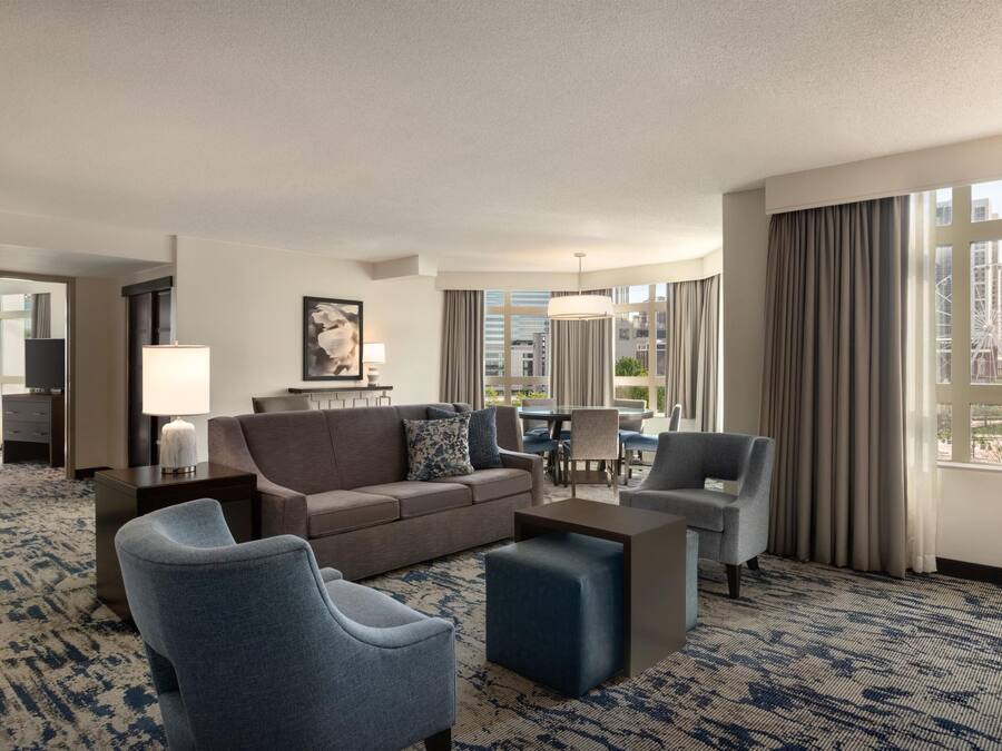 Habitación de hotel con sofá, sillones, una mesa de centro, televisor, lámpara, un cuadro y vistas a la ciudad.