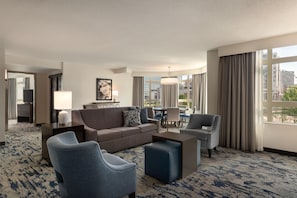 Une chambre d’hôtel avec un canapé, des fauteuils, une table basse, un téléviseur, une lampe, un tableau et une vue sur la ville.