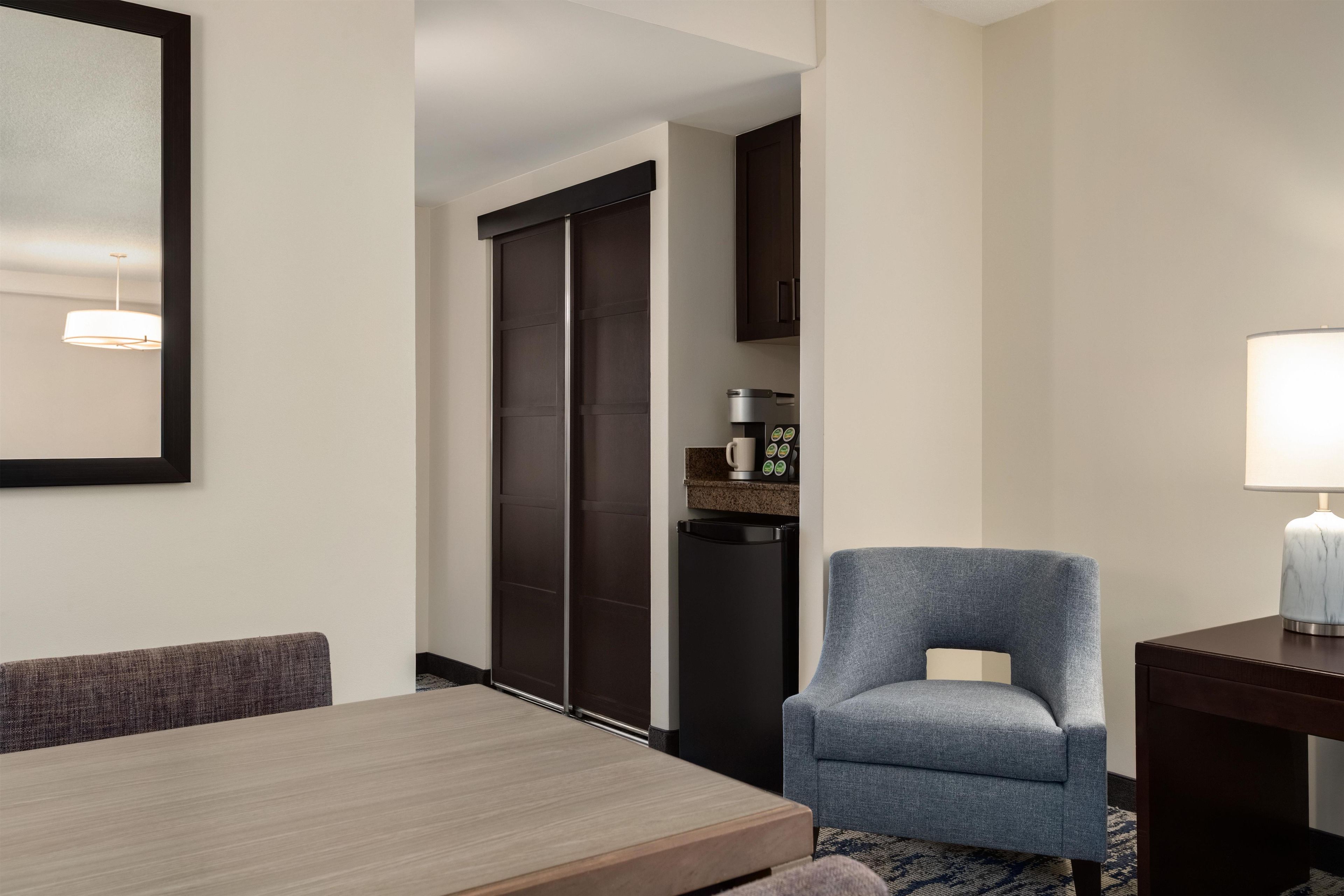 A hotel room with a dining table, a blue armchair, a coffee machine, and a lamp.