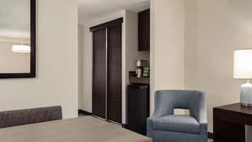 A hotel room with a dining table, a blue armchair, a coffee machine, and a lamp.