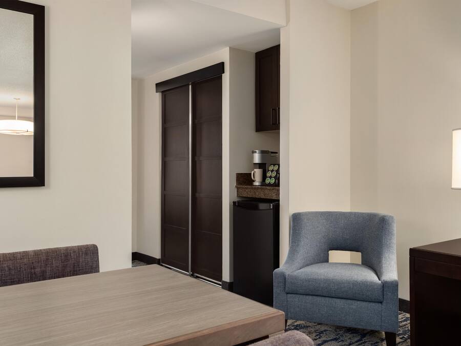 A hotel room with a dining table, a blue armchair, a coffee machine, and a lamp.