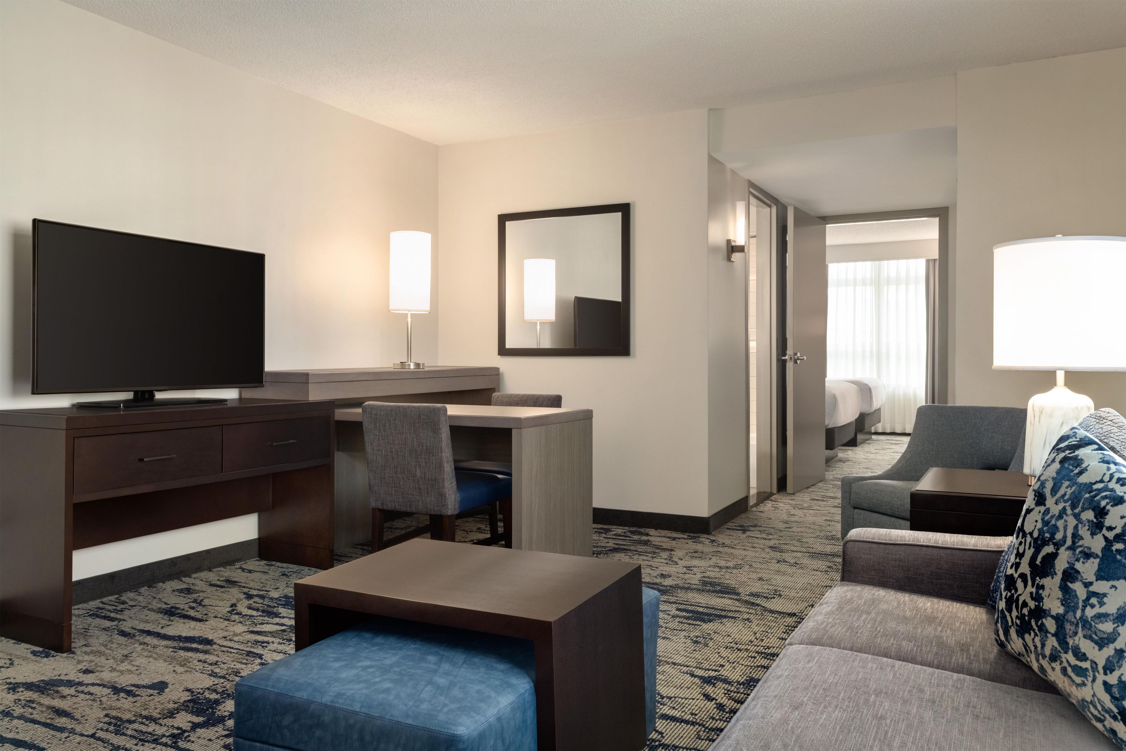 Een hotelkamer met een flatscreen televisie, een bureau met stoel, een bank, een kamerbreed tapijt met dessin en een grote spiegel.
