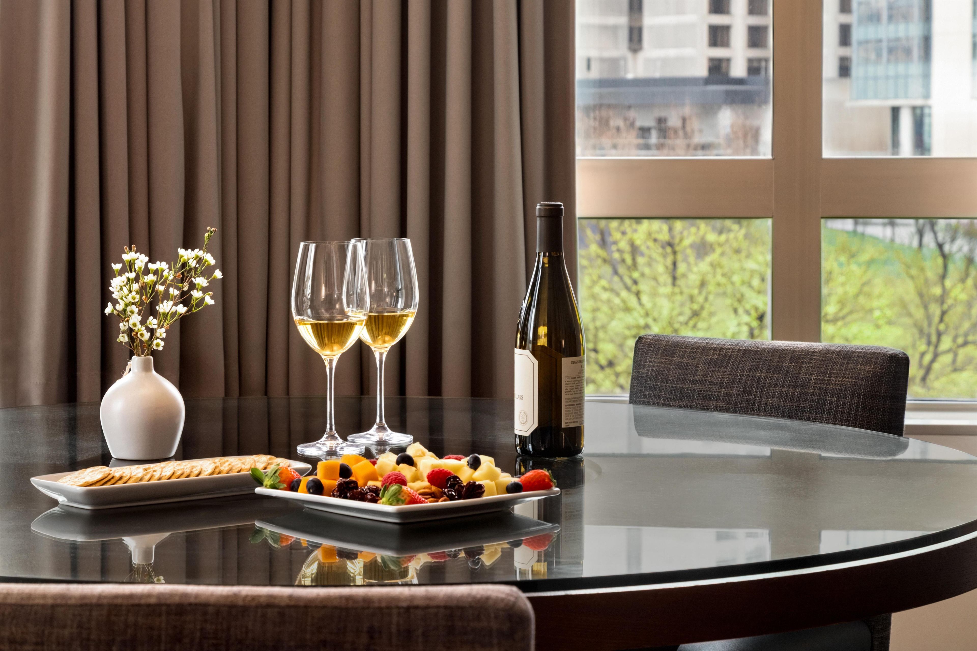 A table set with a bottle of wine, two glasses of white wine, a plate of assorted fruits, and a vase with flowers.