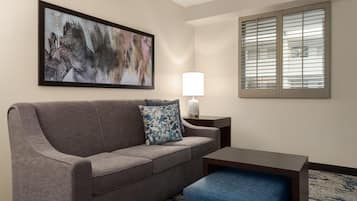 A hotel room with a grey sofa, a blue ottoman, a white lamp, and a painting on the wall.