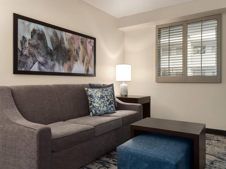 Une chambre d’hôtel avec un canapé gris, un pouf bleu, une lampe blanche et un tableau accroché au mur.