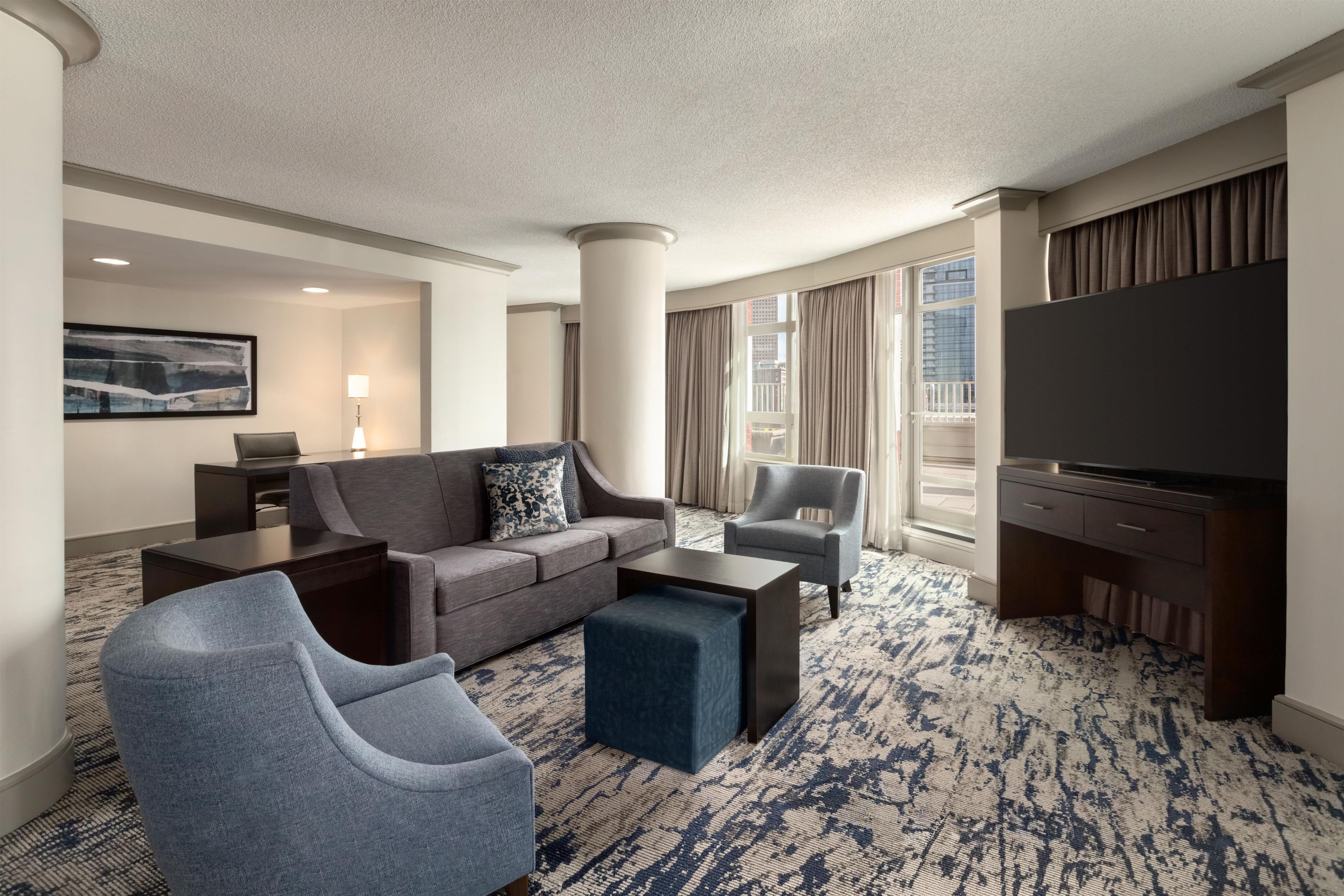 Quarto de hotel com uma televisão de tela grande, um sofá cinza, uma poltrona azul, uma mesa de centro e um tapete estampado.
