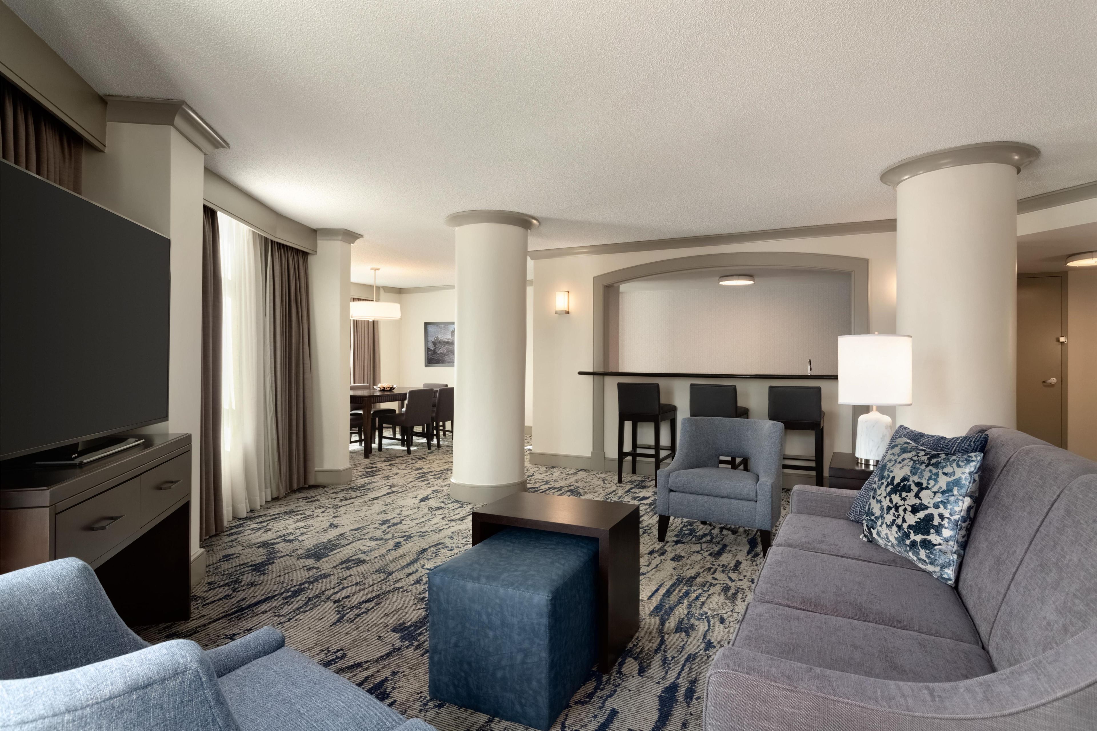 A hotel room with a flat-screen TV, a gray sofa, a blue ottoman, and a dining area with a table and chairs.