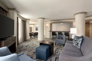 A hotel room with a flat-screen TV, a gray sofa, a blue ottoman, and a dining area with a table and chairs.