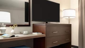Habitación de hotel con televisión de pantalla plana, un escritorio con lámpara, un florero con flores, una taza y un libro.
