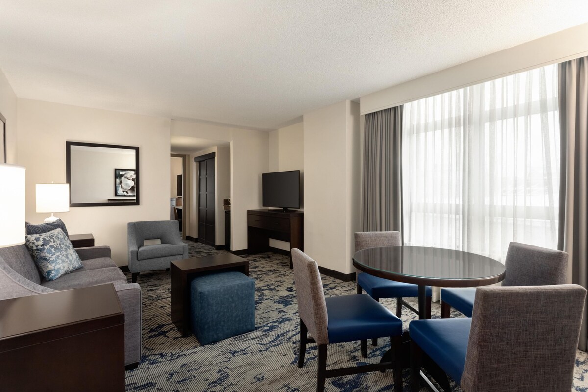 Een hotelkamer met een bank, fauteuil, poef, een eettafel met glazen blad en stoelen, een televisie en een groot raam met gordijnen.