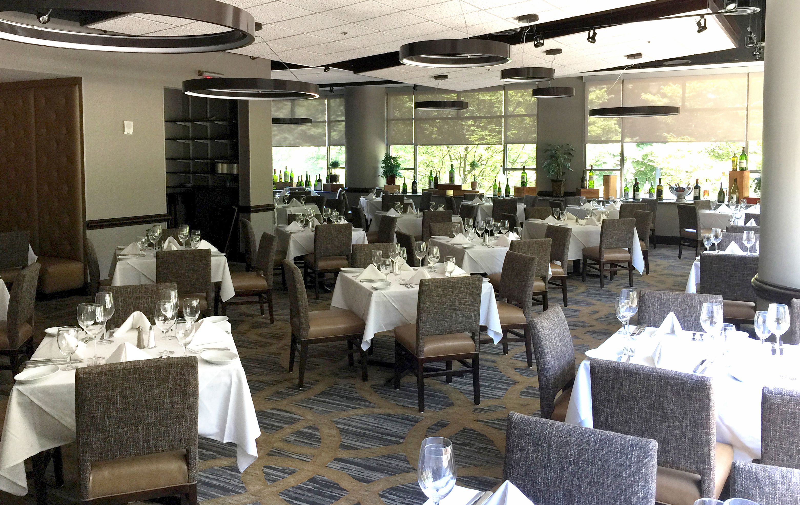 Un vaste espace dédié à la salle à manger, avec plusieurs tables dressées, de grandes fenêtres laissant entrer la lumière naturelle et un sol recouvert de moquette.